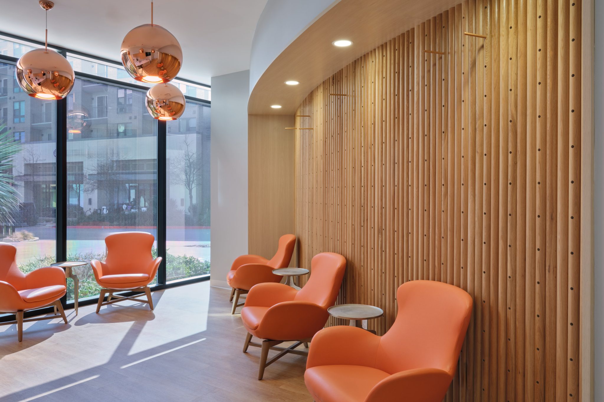 Sunlit lobby with copper pendant lights and orange lounge chairs against a slatted wood wall