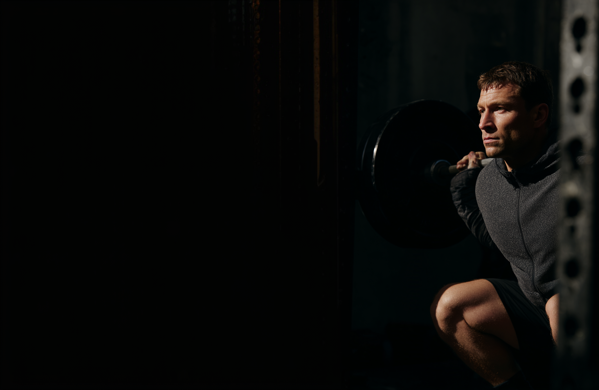 Athletic man crouched with a loaded barbell in a dark gym, lit from the side