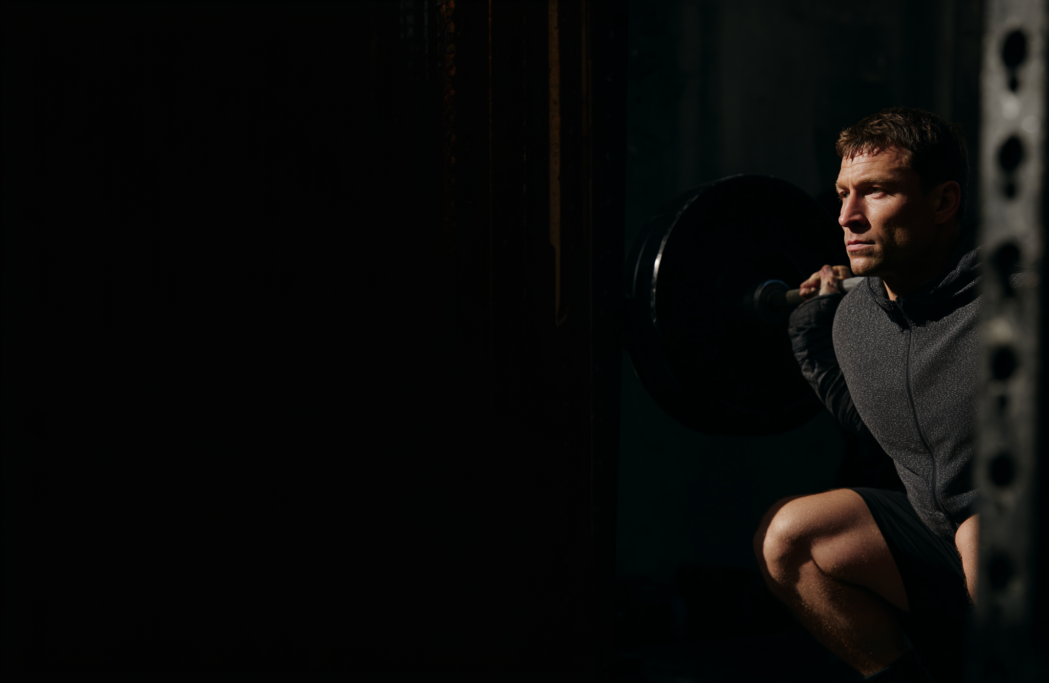 Athletic man crouched with a loaded barbell in a dark gym, lit from the side
