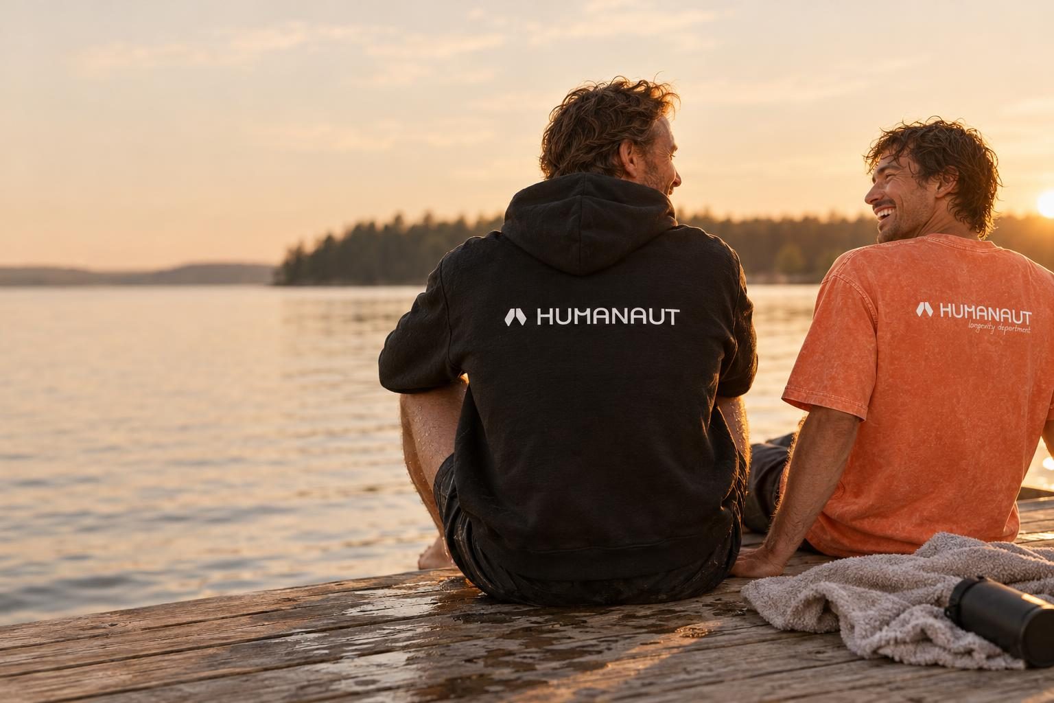 Two friends sitting on a lake dock at sunset, one in a black Humanaut hoodie, one in an orange Humanaut tee