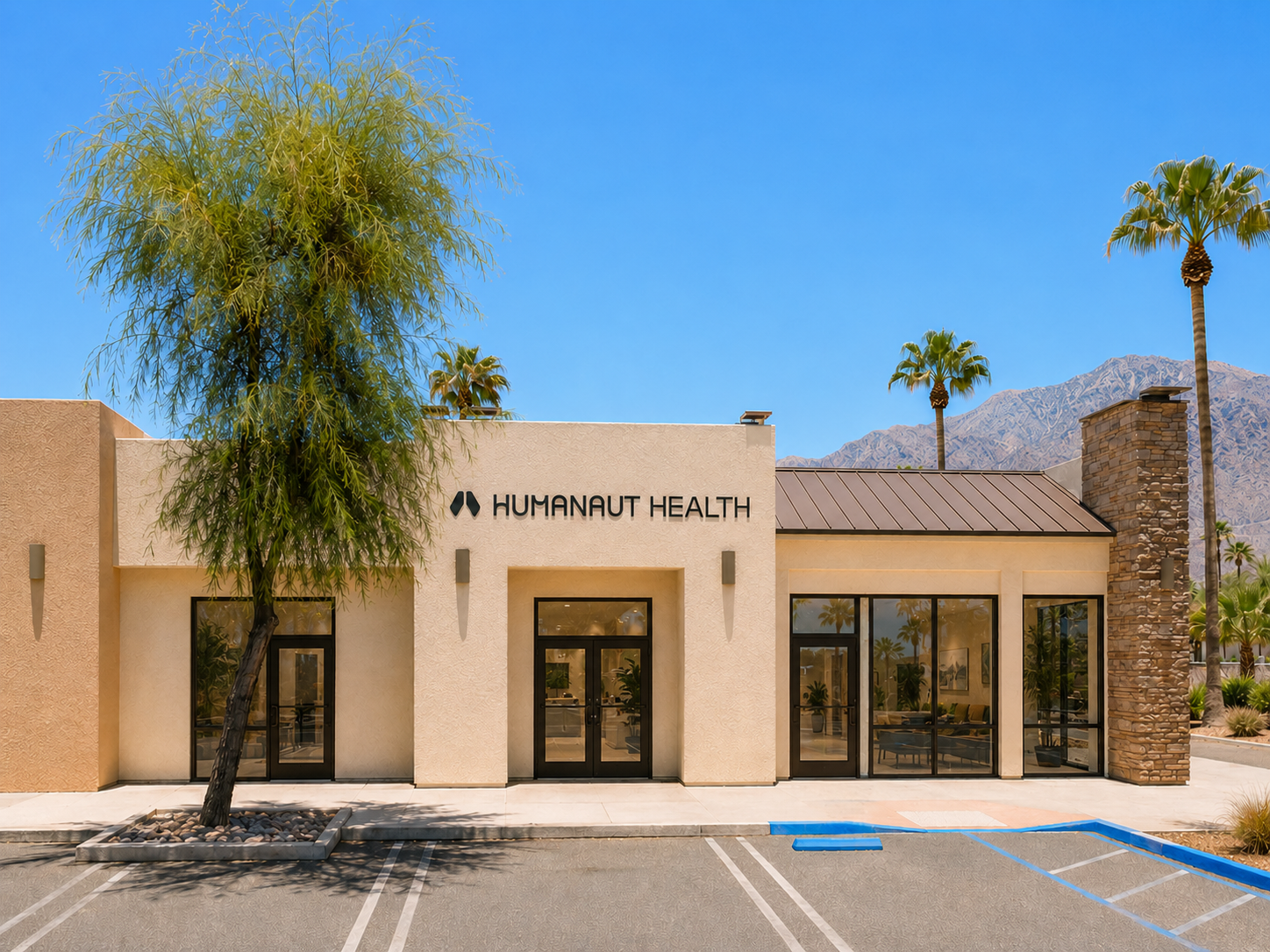Humanaut Health clinic exterior, a single-story tan stucco building with the company name and logo above glass double doors, framed by desert palms and mountains under a clear blue sky.