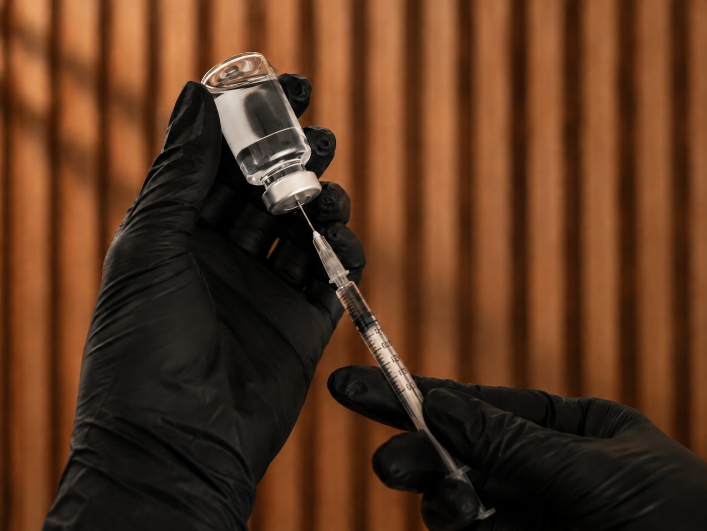 Gloved hands drawing medication from a vial with a slatted wood backdrop